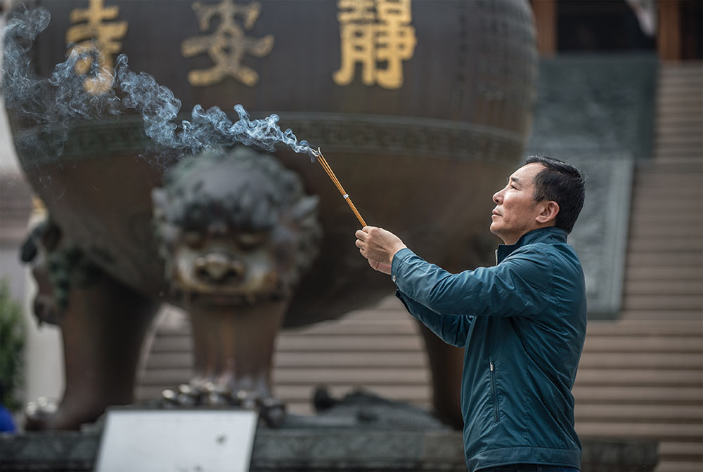 A man waves sticks of incense in the air next to an ancient statue in Jing'an Temple of Shanghai area.