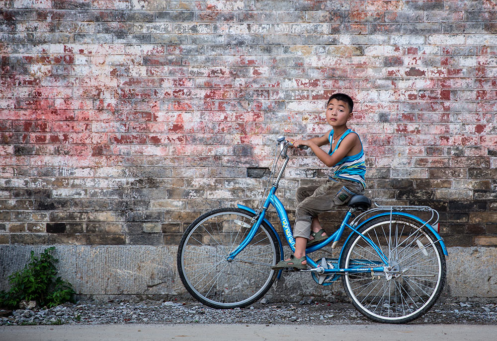 A young boy leans on a blue bicycle next to a brick wall in Jiuxian village in Yangshuo, China.