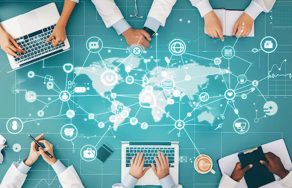 Graphic of a world map with network of heathcare industry icons as nodes. This is overlayed on a table with six doctors hands holding notebooks and computers. 