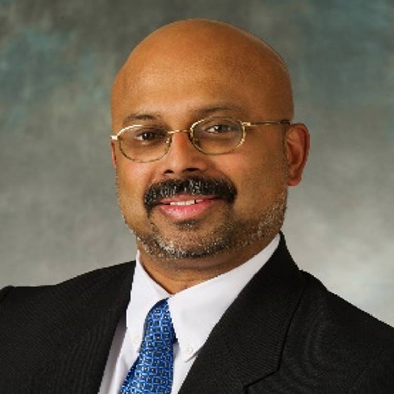 A man with tan skin, wearing glasses, has a mustache and beard, dark blazer, dress shirt and tie