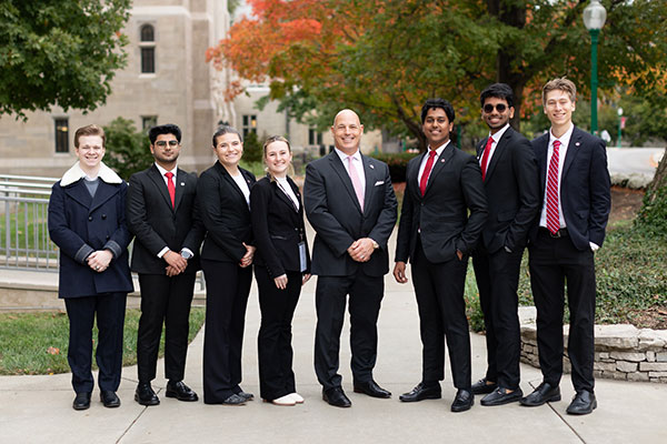 Group of seven students and one professor, all in formal attire, standing and smiling outdoors on the Kelley School of Business campus in autumn.