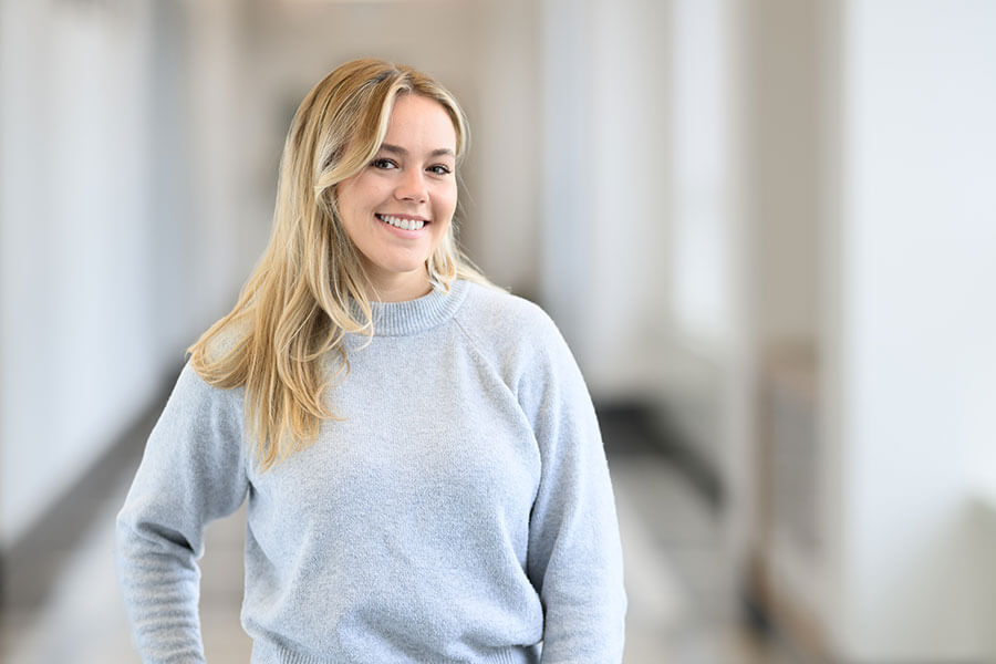 A smiling student wearing a gray sweater stands in the hallway that connects the Hodge Hall Undergraduate Center and the William J. Godfrey Graduate and Executive Education Center buildings at the Kelley School of Business.