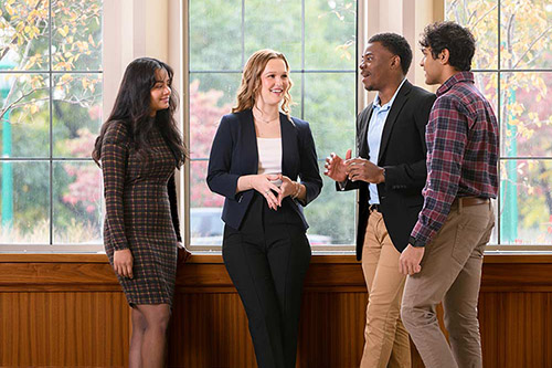 A group of four undergraduate students from the Kelley School of Business chat together in front of a window that overlooks the campus. 