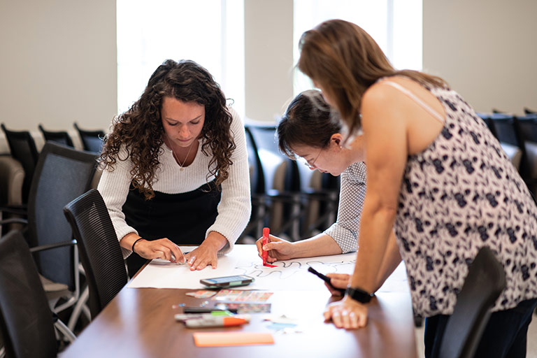 A group of Kelley School of Business instructors work together on a project. They are gathered around a table and are writing on a large sheet of paper. 