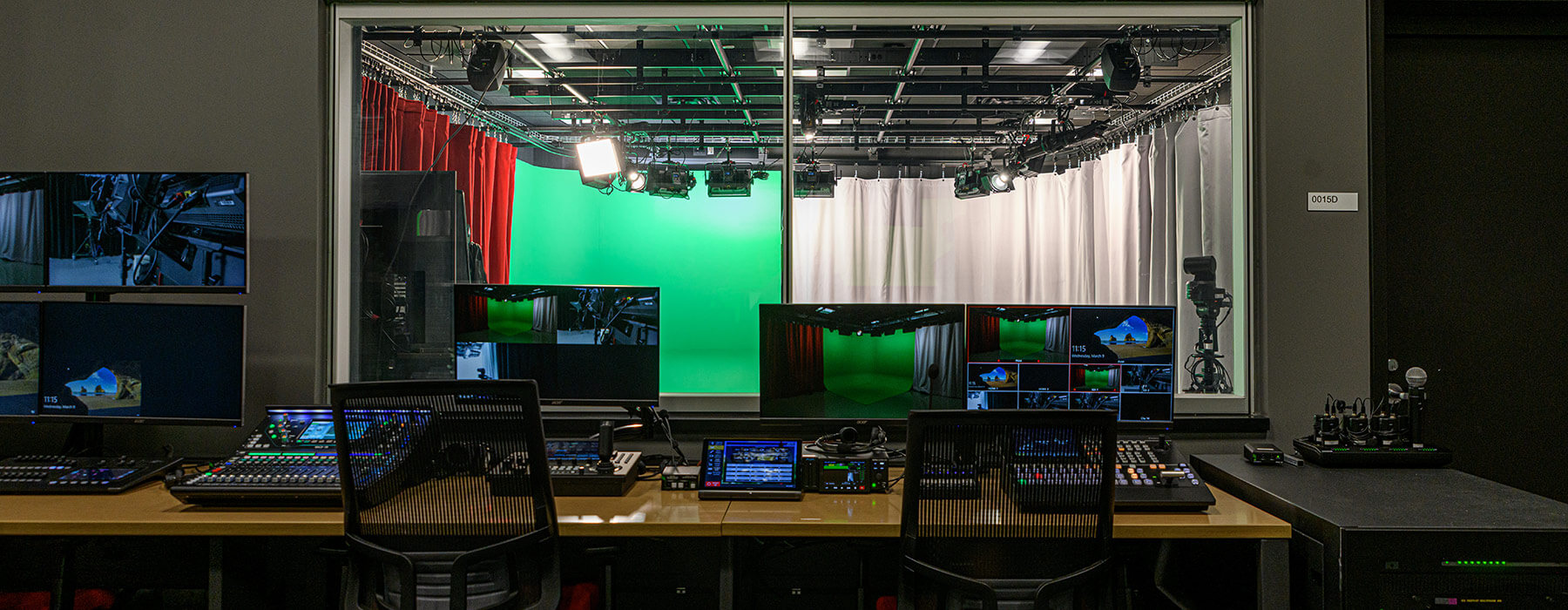 Control room of Jellison Studios shows computer monitor displaying several camera angles of one of the Black Box Studios. Through the control room window, the view of the studio room shows a partial red curtain, a green screen, and a white curtain. Lights and cameras are mounted in the ceiling.