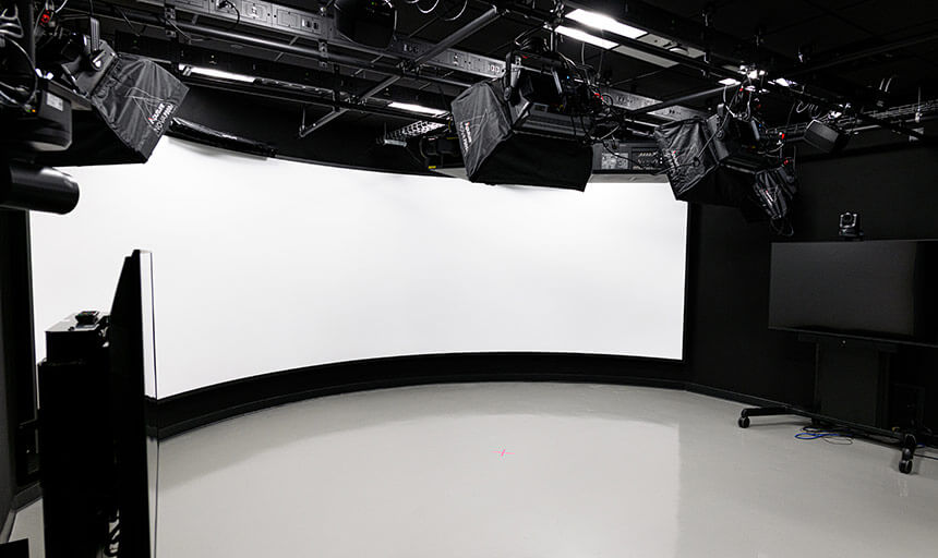 An interior of a projection studio shows the white curved projection wall. The wall is surrounded by video equipment and lights are mounted above in the ceiling.