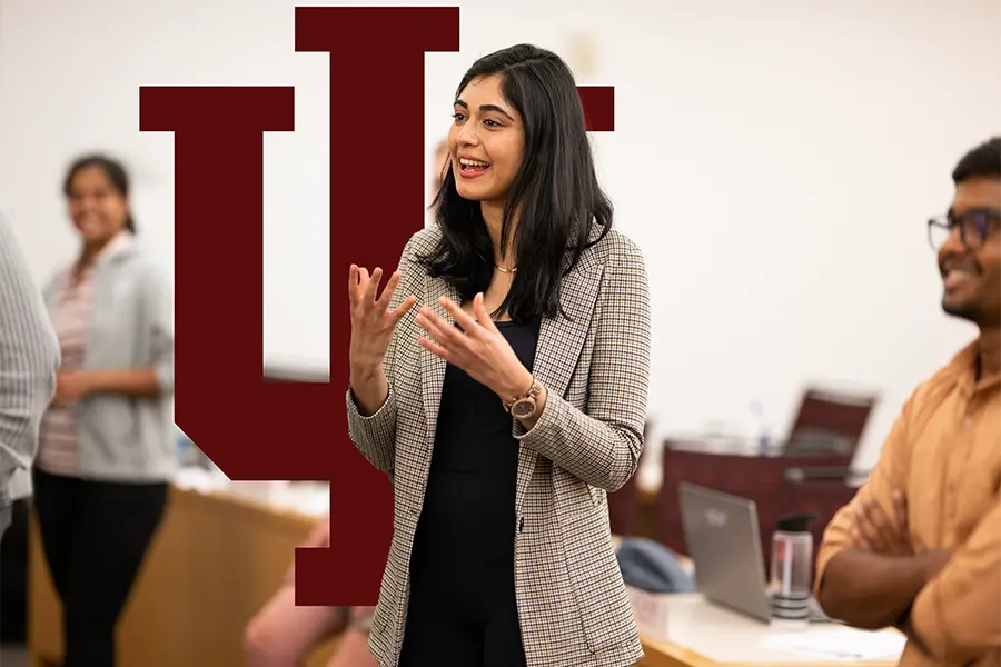 An MBA student gesturing while speaking in a classroom setting with the Indiana University trident prominently displayed in the background behind the student. Other attendees are visible, smiling and listening attentively.