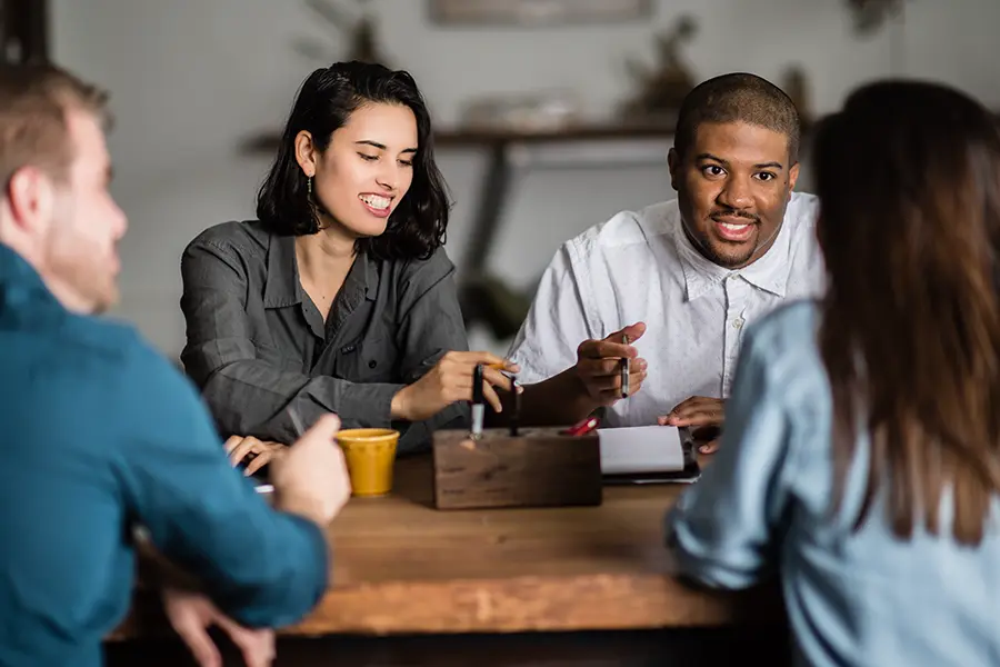 Four people engaged in a lively meeting around a wooden table, with documents and a digital tablet visible.