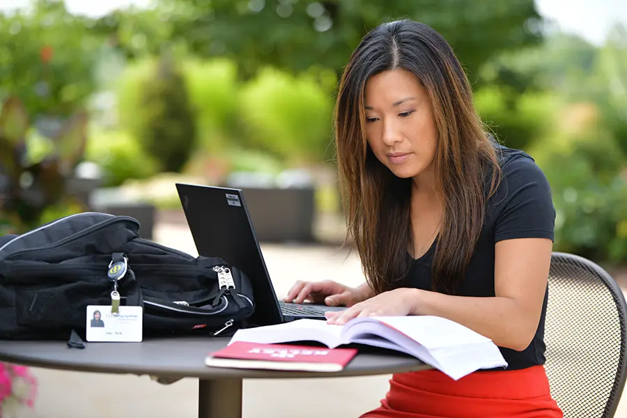 Individual studying outdoors in a green space with a laptop and textbooks on a table, with a backpack and identification badge visible.