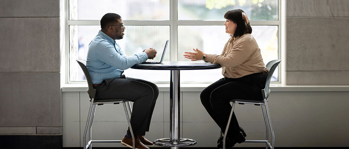 Two individuals are seated at a small table, engaged in a conversation with a laptop open between them. They are in a bright room with large windows.