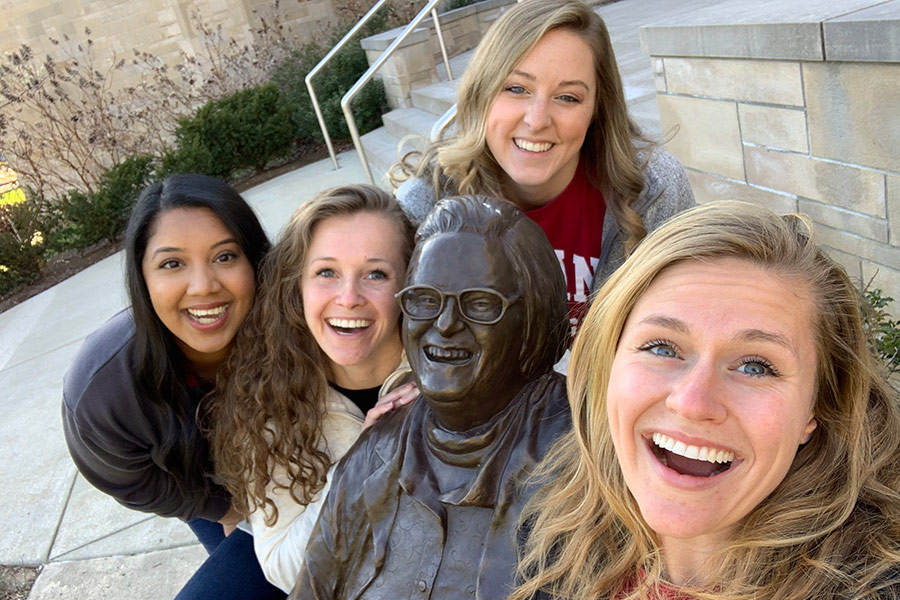 A group of four graduate business students pose for a photo around a statue of Nobel Prize-winner Elinor Ostrom on the Indiana University Bloomington campus.