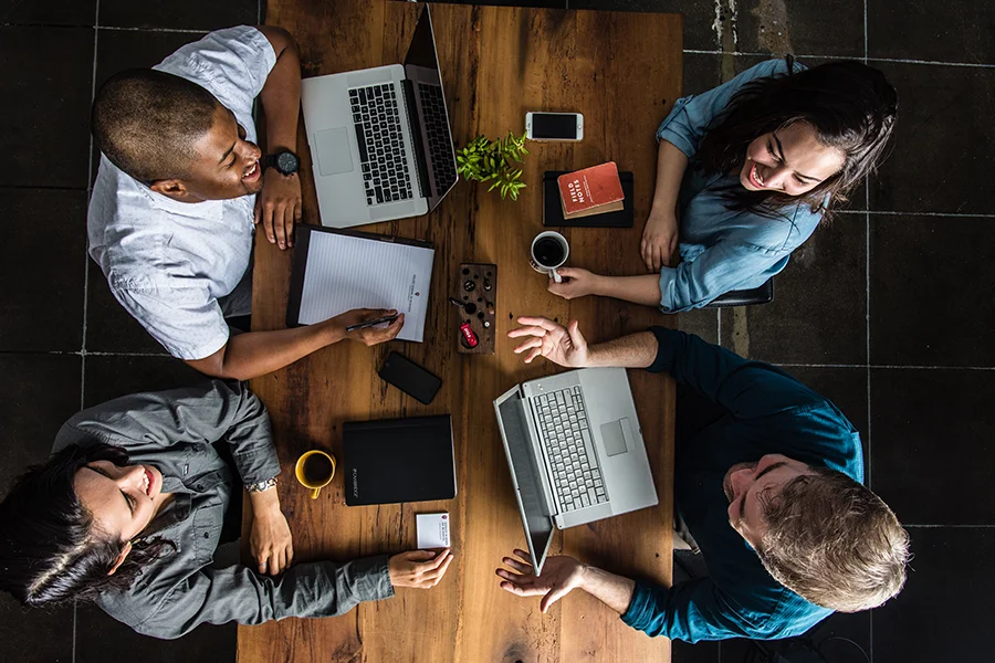 A group of MBA students talking at a table with laptops, notes, and coffee.