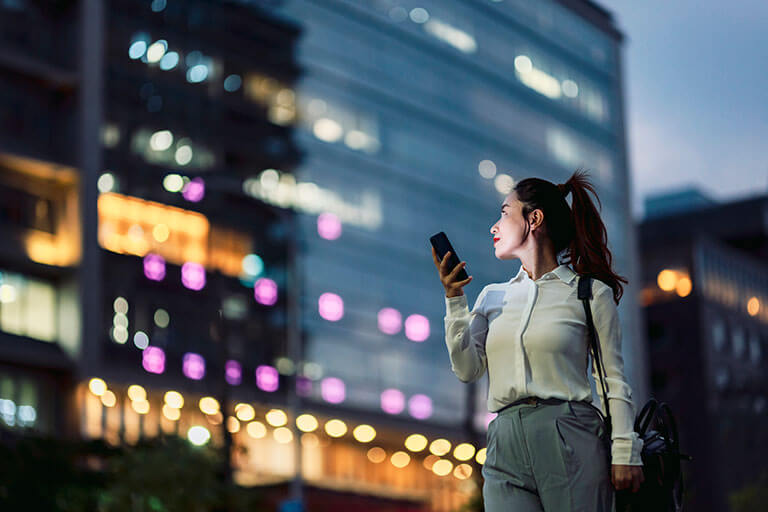 A business woman holds a mobile and looks up at building that has many office lights on. 