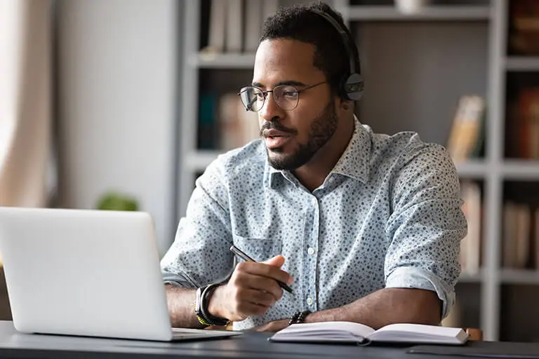 A person in a dotted shirt, wearing headphones, sits at a desk with a laptop and notebook, focused on work in a cozy setting.