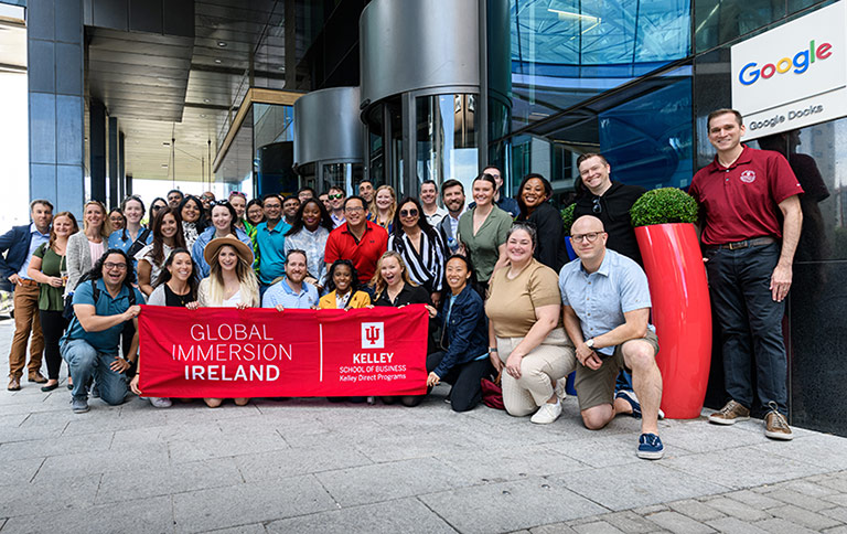 A group of more than 30 Kelley Direct Online MBA students gather in front of Google in Dublin, Ireland. They are holding a red banner that reads Global Immersion Ireland, Kelley School of Business Kelley Direct Program.